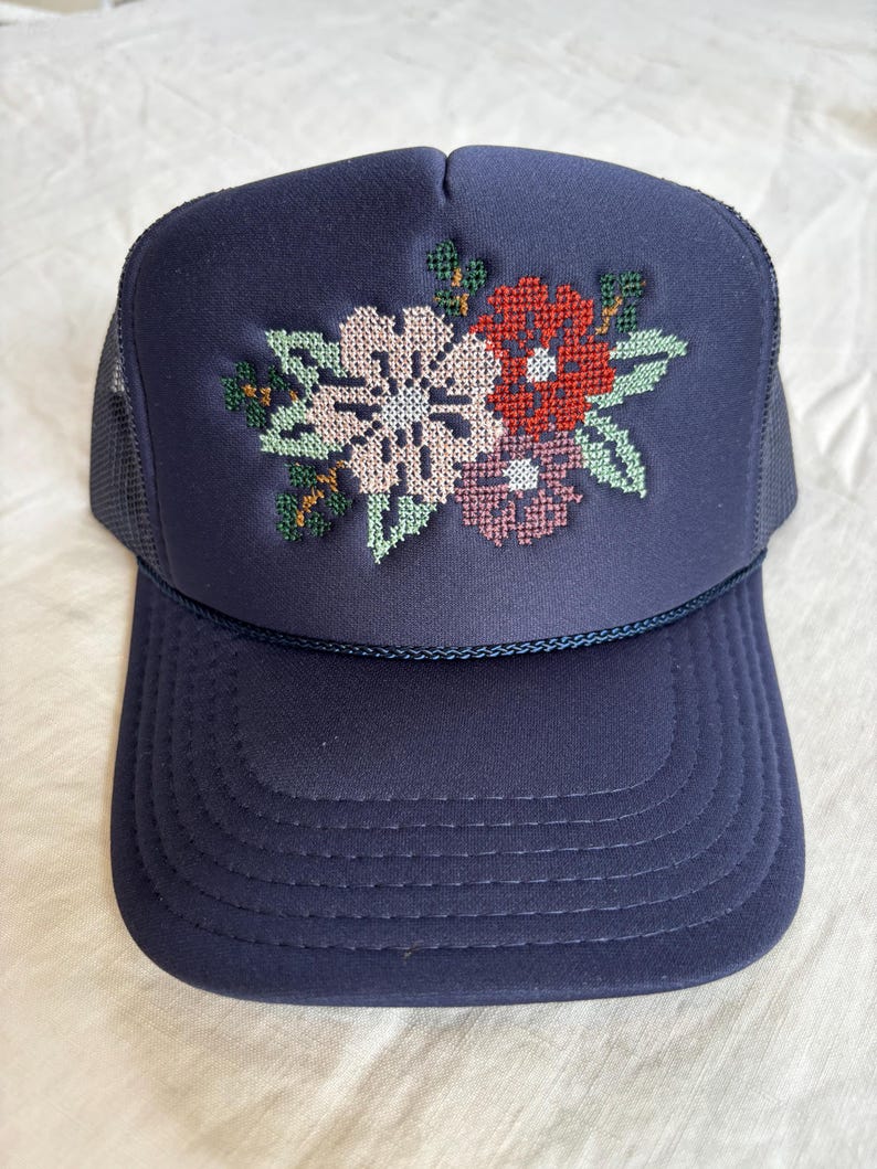May include: Navy blue trucker hat featuring a floral cross-stitch design. The embroidered flowers include white, red, and pink blooms with green leaves. The hat has a mesh back and a curved peak.