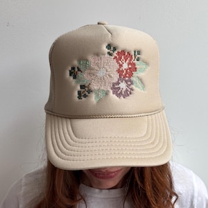 May include: A beige trucker hat featuring a cross-stitch style floral embroidery on the front panel. The design includes light pink, red, and purple flowers with green leaves. The hat has a matching beige brim, a braided cord, and mesh back.