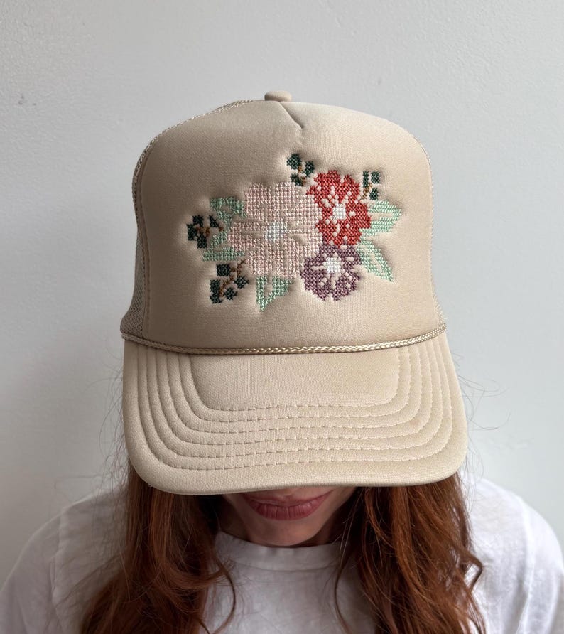 May include: A beige trucker hat featuring a cross-stitch style floral embroidery on the front panel. The design includes light pink, red, and purple flowers with green leaves. The hat has a matching beige brim, a braided cord, and mesh back.