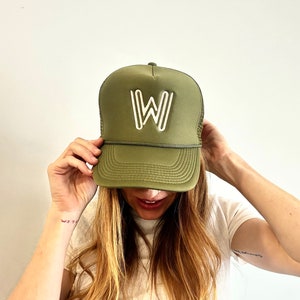 May include: A green trucker hat with a white embroidered "W" on the front. The hat is worn by a person with long brown hair.
