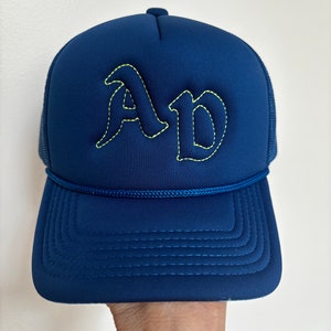 May include: A navy blue trucker hat featuring the letters AD embroidered in a bold, gothic-style font with bright yellow stitching. The cap includes a matching navy blue cord trim across the base of the crown and a structured front panel.