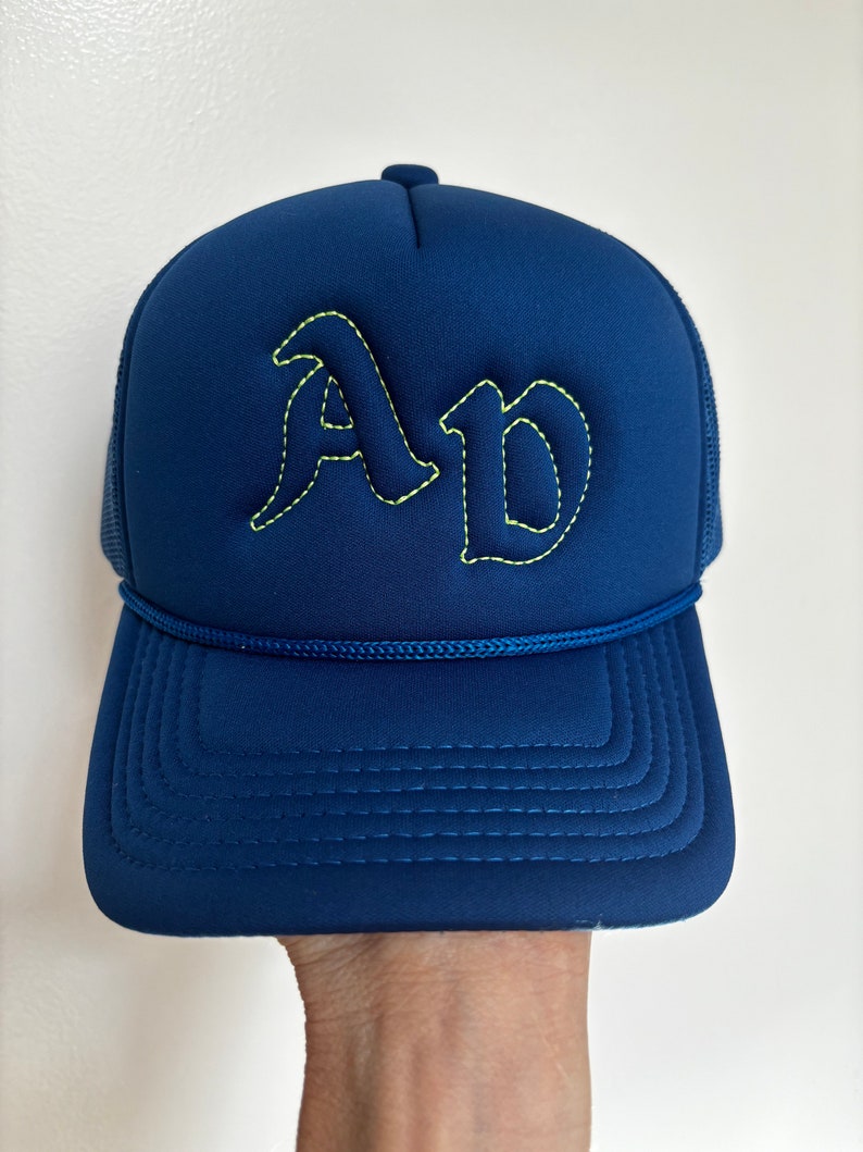 May include: A navy blue trucker hat featuring the letters AD embroidered in a bold, gothic-style font with bright yellow stitching. The cap includes a matching navy blue cord trim across the base of the crown and a structured front panel.