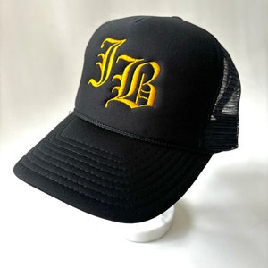 May include: A black trucker hat featuring a structured front panel with the letters JB embroidered in a bold yellow gothic font. The cap includes a black mesh back, a curved brim with decorative stitching, and a thin black cord across the base.