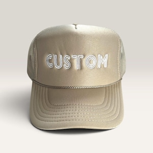May include: A beige trucker hat featuring a mesh back and a solid front panel with the word CUSTOM embroidered in white thread. The cap includes a decorative braided cord across the base of the crown and a curved brim with stitched detailing.