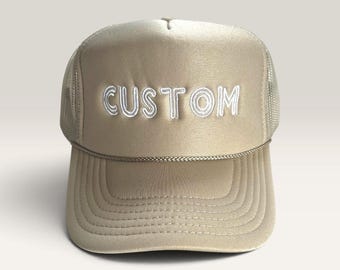May include: A beige trucker hat featuring a mesh back and a solid front panel with the word CUSTOM embroidered in white thread. The cap includes a decorative braided cord across the base of the crown and a curved brim with stitched detailing.