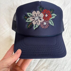 May include: A navy blue trucker hat featuring a cross-stitch floral embroidery design on the front panel. The cap includes a matching navy blue cord trim across the base of the crown and a mesh back panel for ventilation.