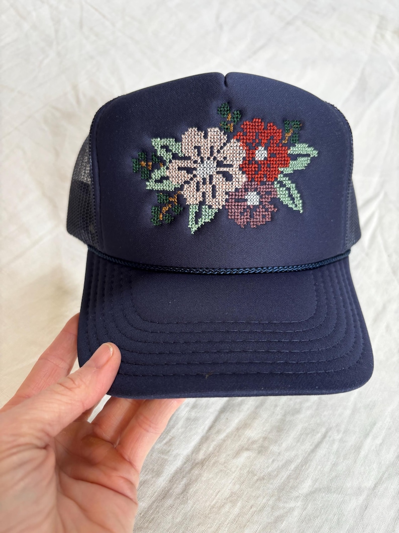 May include: A navy blue trucker hat featuring a cross-stitch floral embroidery design on the front panel. The cap includes a matching navy blue cord trim across the base of the crown and a mesh back panel for ventilation.