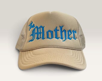 Könnte beinhalten: Eine beige Trucker-Cap mit Netzrückseite und einem Vorderteil aus Schaumstoff. Das Wort "The Mother" ist in leuchtend blauer, gotischer Schrift auf die Vorderseite gestickt. Eine dünne, dekorative Kordel verläuft horizontal über den unteren Rand.
