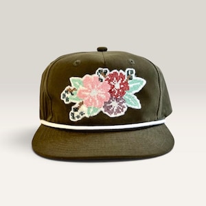 May include: An olive green baseball cap featuring a white cord trim along the base of the crown. The front panel displays a cross-stitch style embroidered floral patch with pink, red, and dark purple flowers surrounded by light green leaves.