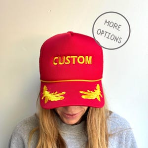 May include: Red trucker hat with a yellow "CUSTOM" embroidered on the front and two yellow embroidered wings on the bill.