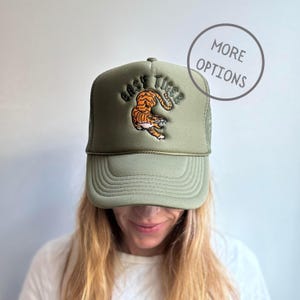 May include: Olive green trucker hat with a mesh back and a curved brim. The hat features an embroidered tiger design in orange and black, with the words "EASY TIGER" above it. A casual accessory.