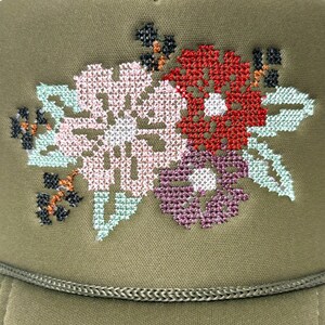 May include: An olive green trucker hat with a floral cross-stitch design. The embroidered flowers are in shades of pink, red, purple, and light green. The hat has a braided trim along the bottom edge.