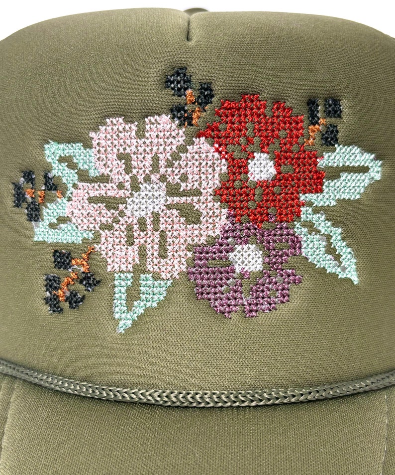 May include: An olive green trucker hat with a floral cross-stitch design. The embroidered flowers are in shades of pink, red, purple, and light green. The hat has a braided trim along the bottom edge.