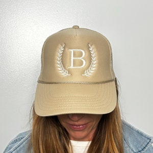 May include: A beige trucker hat featuring a white embroidered letter B centered between two white laurel leaf branches. The cap includes a matching beige brim, a decorative braided cord across the base of the front panel, and a mesh back section.