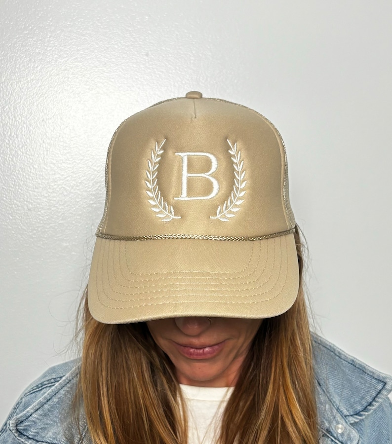 May include: A beige trucker hat featuring a white embroidered letter B centered between two white laurel leaf branches. The cap includes a matching beige brim, a decorative braided cord across the base of the front panel, and a mesh back section.