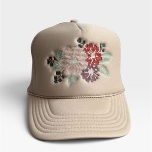 May include: A beige trucker hat featuring a cross-stitch style floral embroidery on the front panel. The design includes light pink, red, purple, and green thread colors. The cap has a structured front, a curved brim, and a matching beige cord trim.