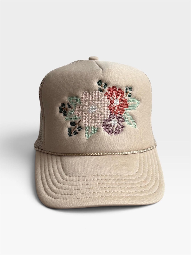 May include: A beige trucker hat featuring a cross-stitch style floral embroidery on the front panel. The design includes light pink, red, purple, and green thread colors. The cap has a structured front, a curved brim, and a matching beige cord trim.