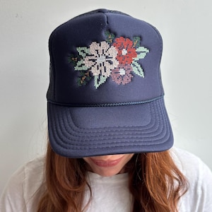 May include: A navy blue trucker hat featuring a structured front panel with a cross-stitch style floral embroidery. The design displays a cluster of flowers in shades of light pink, red, and dark purple, accented with green leaves. The hat has a mesh back.