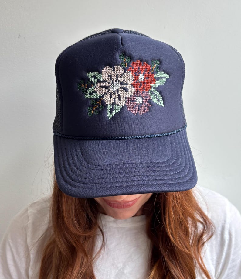May include: A navy blue trucker hat featuring a structured front panel with a cross-stitch style floral embroidery. The design displays a cluster of flowers in shades of light pink, red, and dark purple, accented with green leaves. The hat has a mesh back.