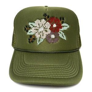May include: An olive green trucker hat with a cross-stitch floral design. The embroidered flowers are white, burgundy, and purple with green leaves. The hat has a curved peak and a mesh back.