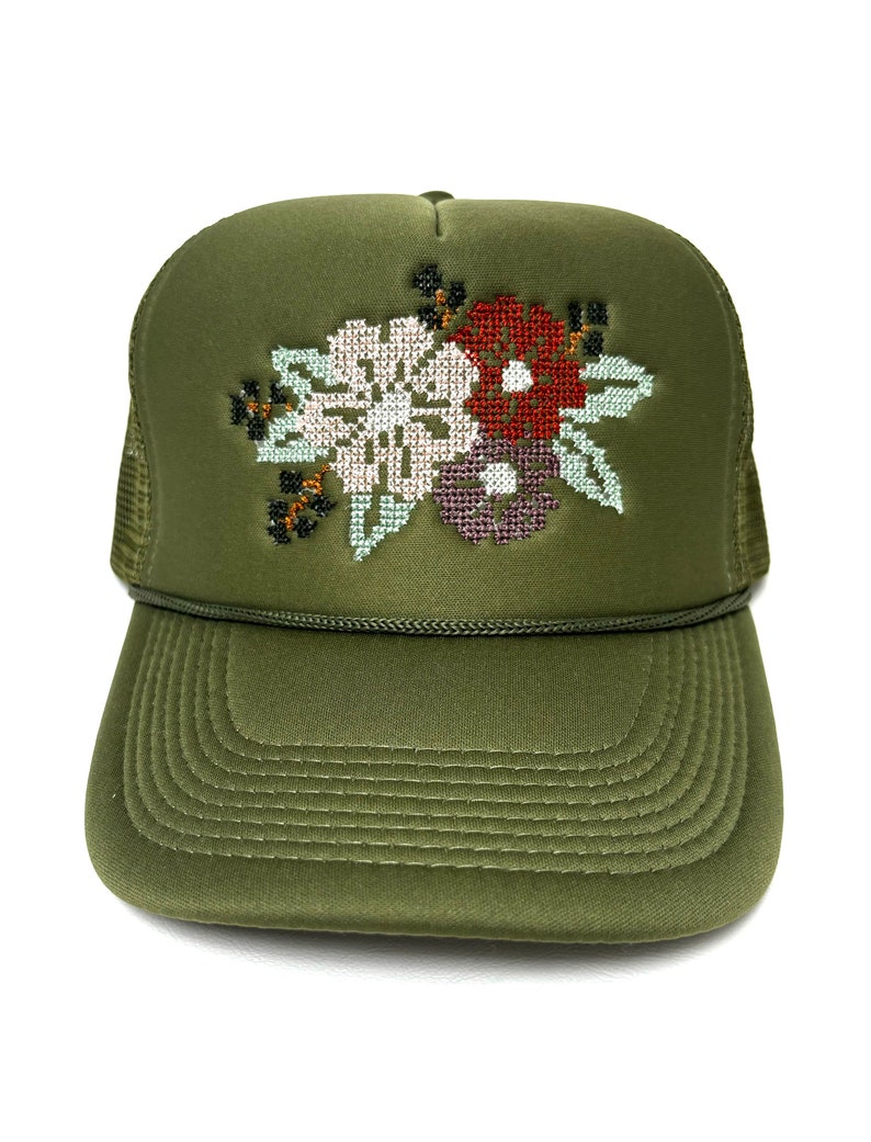 May include: An olive green trucker hat with a cross-stitch floral design. The embroidered flowers are white, burgundy, and purple with green leaves. The hat has a curved peak and a mesh back.