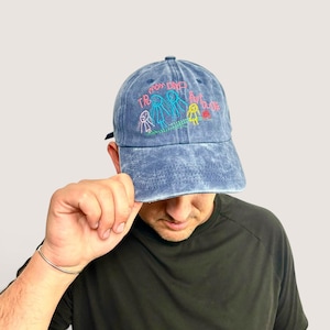 May include: A blue acid-wash baseball cap featuring embroidered stick figure drawings in pink, blue, yellow, and green thread. The embroidery includes the text "MON DAD" and "AVOUE" in pink, depicting a family scene with three figures.
