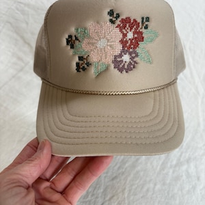May include: A tan trucker hat with a floral cross-stitch design. The hat has a beige front panel with a stitched flower pattern in pink, red, purple, and green. The mesh back and brim are tan, with a gold rope detail.