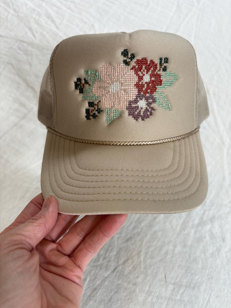 May include: A tan trucker hat with a floral cross-stitch design. The hat has a beige front panel with a stitched flower pattern in pink, red, purple, and green. The mesh back and brim are tan, with a gold rope detail.