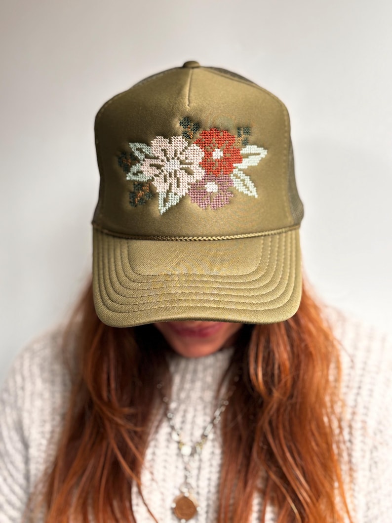 May include: Olive green trucker cap featuring a floral cross-stitch design in pink, red, and white. The cap has a mesh back and a curved peak. A fashionable accessory for casual wear.