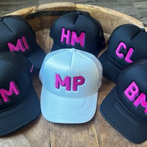 May include: A collection of six trucker hats featuring mesh backs and foam fronts. Five hats are black with pink embroidered initials MI, HM, CL, JM, and BM. One central hat is white with pink embroidered initials MP. Each hat has a structured crown.