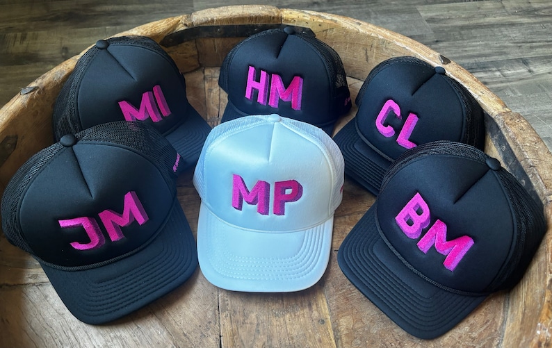 May include: A collection of six trucker hats featuring mesh backs and foam fronts. Five hats are black with pink embroidered initials MI, HM, CL, JM, and BM. One central hat is white with pink embroidered initials MP. Each hat has a structured crown.