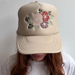 May include: A beige trucker cap featuring a floral cross-stitch design in red, pink, purple, and green. The hat has a mesh back and a curved peak. A fashionable accessory.