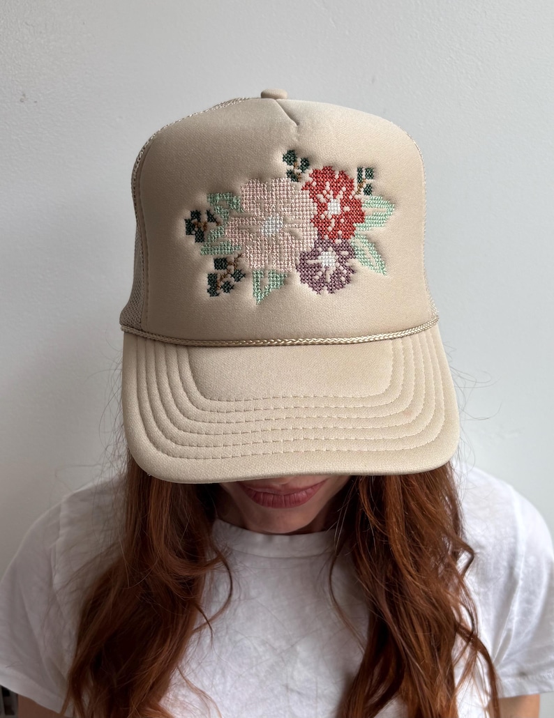 May include: A beige trucker cap featuring a floral cross-stitch design in red, pink, purple, and green. The hat has a mesh back and a curved peak. A fashionable accessory.