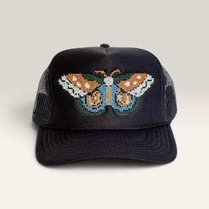 May include: A black trucker hat featuring a cross-stitch style embroidered butterfly on the front panel. The butterfly design incorporates shades of orange, light blue, white, and dark green. The hat has a solid black brim and mesh back panels.