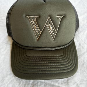 May include: An olive green trucker hat featuring a foam front panel with an embroidered letter W in a light tan thread. The letter W has a decorative pattern within its strokes. The hat includes a matching olive green cord across the brim base.