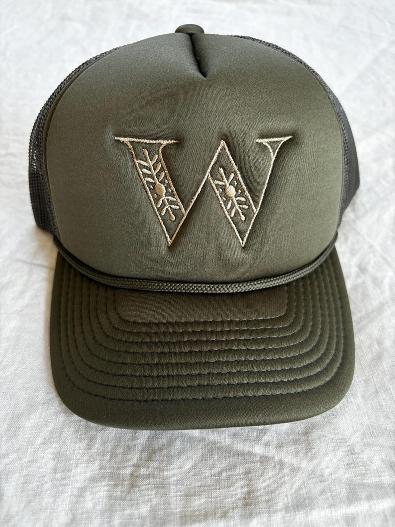 May include: An olive green trucker hat featuring a foam front panel with an embroidered letter W in a light tan thread. The letter W has a decorative pattern within its strokes. The hat includes a matching olive green cord across the brim base.