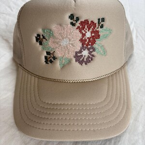 May include: A beige trucker hat featuring a cross-stitch floral embroidery design on the front panel. The embroidery includes light pink, dark red, and purple flowers with green leaves and black accents. A thin gold cord trim sits above the brim.