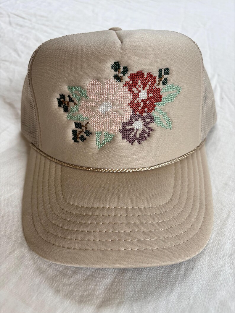 May include: A beige trucker hat featuring a cross-stitch floral embroidery design on the front panel. The embroidery includes light pink, dark red, and purple flowers with green leaves and black accents. A thin gold cord trim sits above the brim.