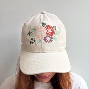 May include: A cream-colored baseball cap featuring a cross-stitch floral embroidery design on the front panel. The embroidery depicts a cluster of flowers in shades of light pink, deep red, and purple, accented with green leaves and dark stems.
