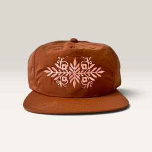 May include: A rust-colored baseball cap featuring a symmetrical, light pink embroidered floral design on the front panel. The cap has a flat brim with visible stitching and a structured crown with a small button at the top center.