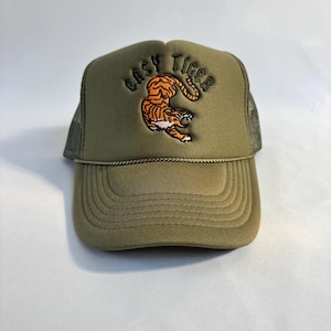 May include: An olive green trucker hat featuring an embroidered orange and black tiger graphic in the center. Above the tiger, the text "EASY TIGER" is embroidered in black. The hat includes a matching olive green brim and a braided cord detail.