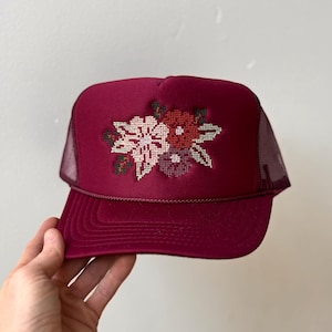 May include: A deep burgundy trucker hat featuring a structured foam front panel with a cross-stitch floral embroidery design in shades of pink, red, and dark green. The cap includes a matching burgundy mesh back and a braided cord across the brim.