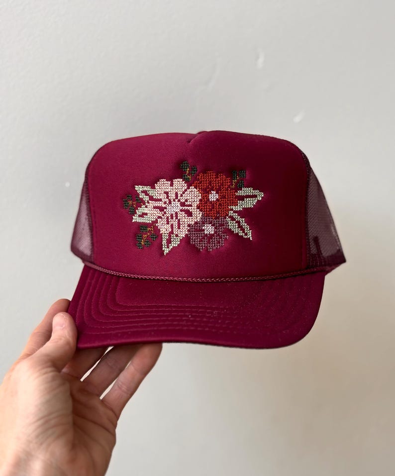 May include: A deep burgundy trucker hat featuring a structured foam front panel with a cross-stitch floral embroidery design in shades of pink, red, and dark green. The cap includes a matching burgundy mesh back and a braided cord across the brim.