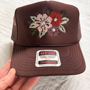 May include: A brown trucker hat featuring a cross-stitched floral design in shades of pink, red, and purple on the front foam panel. The hat includes a mesh back, a curved brim, and a white sticker with the text OTTO WORLD FAMOUS FOAM FRONT OSFM.
