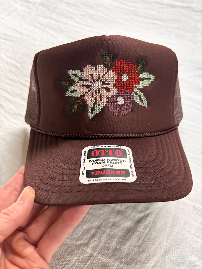 May include: A brown trucker hat featuring a cross-stitched floral design in shades of pink, red, and purple on the front foam panel. The hat includes a mesh back, a curved brim, and a white sticker with the text OTTO WORLD FAMOUS FOAM FRONT OSFM.