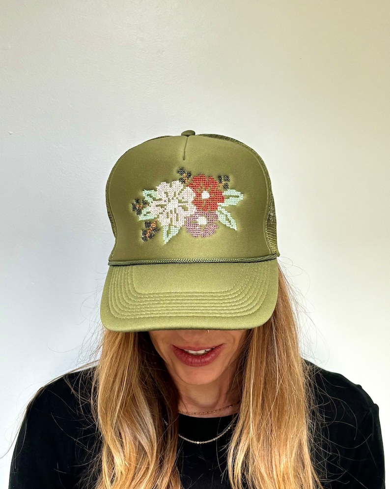 May include: An olive green trucker hat featuring a cross-stitch style floral embroidery on the front panel. The design includes white, red, and light pink flowers with green leaves. The cap has a structured front, a curved brim, and mesh back.