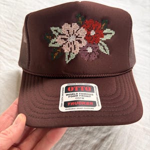May include: A brown trucker hat with a floral cross-stitch design in pink, red, and purple. The hat features a foam front, mesh back, and the text "OTTO TRUCKER" on a tag. OSFM (One Size Fits Most).