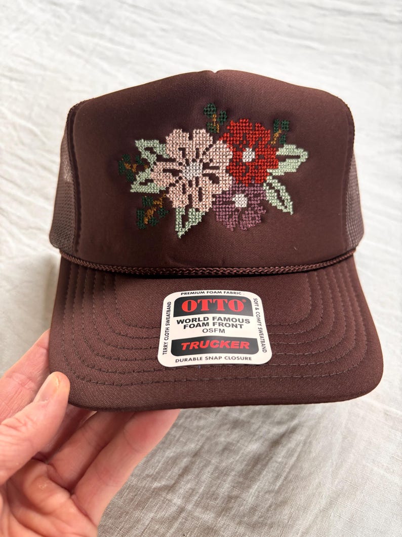 May include: A brown trucker hat with a floral cross-stitch design in pink, red, and purple. The hat features a foam front, mesh back, and the text "OTTO TRUCKER" on a tag. OSFM (One Size Fits Most).