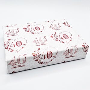 May include: A rectangular gift box wrapped in white paper with a repeating pattern. The pattern features the number "40" in burgundy, along with the words "Wedding Anniversary" and floral heart designs.