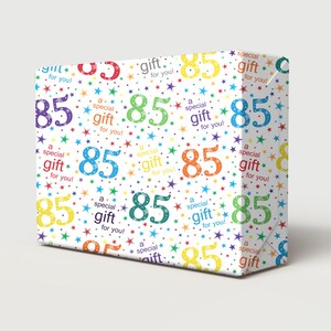Puede incluir: Caja de regalo envuelta con un patrón colorido del número 85, estrellas y la frase "¡un regalo especial para ti!" sobre un fondo blanco. El papel de regalo tiene un tema de cumpleaños.